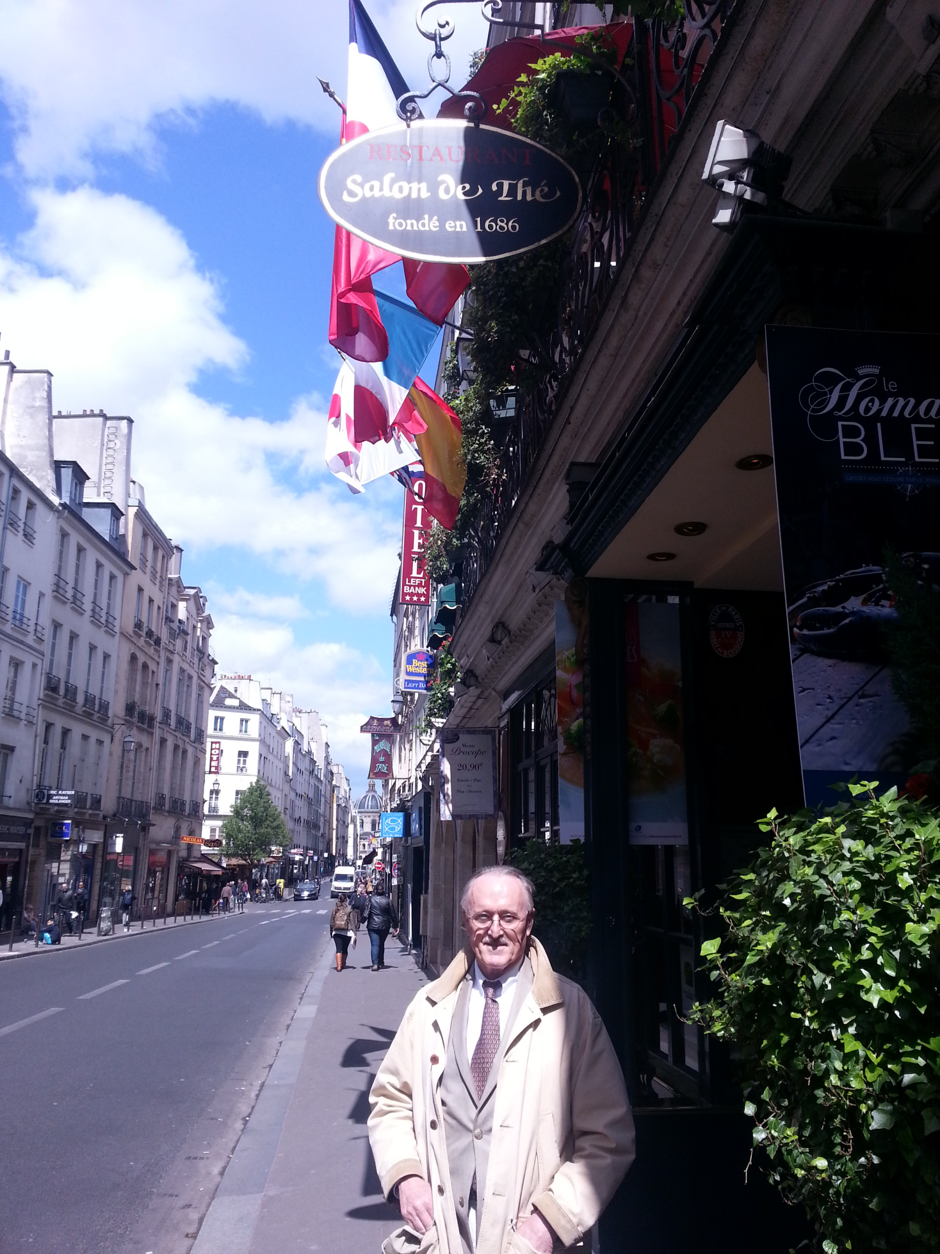 A man in a light coat stands outside the historic Salon de Thé, with French flags displayed above. The charming Parisian street is lined with cafes and shops.