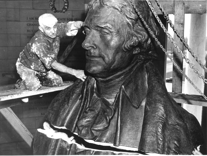 A sculptor carefully works on a large bronze bust of Thomas Jefferson, balancing on scaffolding in front of a stone wall inscribed with text.