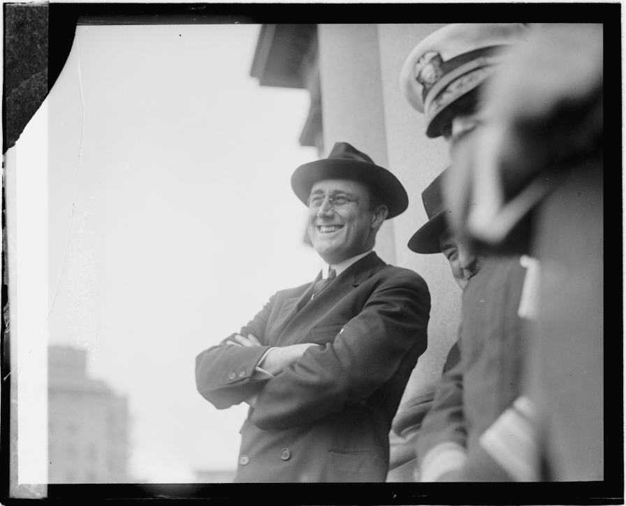 Smiling man in a suit and hat stands with arms crossed, engaged in conversation with two others. Image captures a moment of camaraderie.
