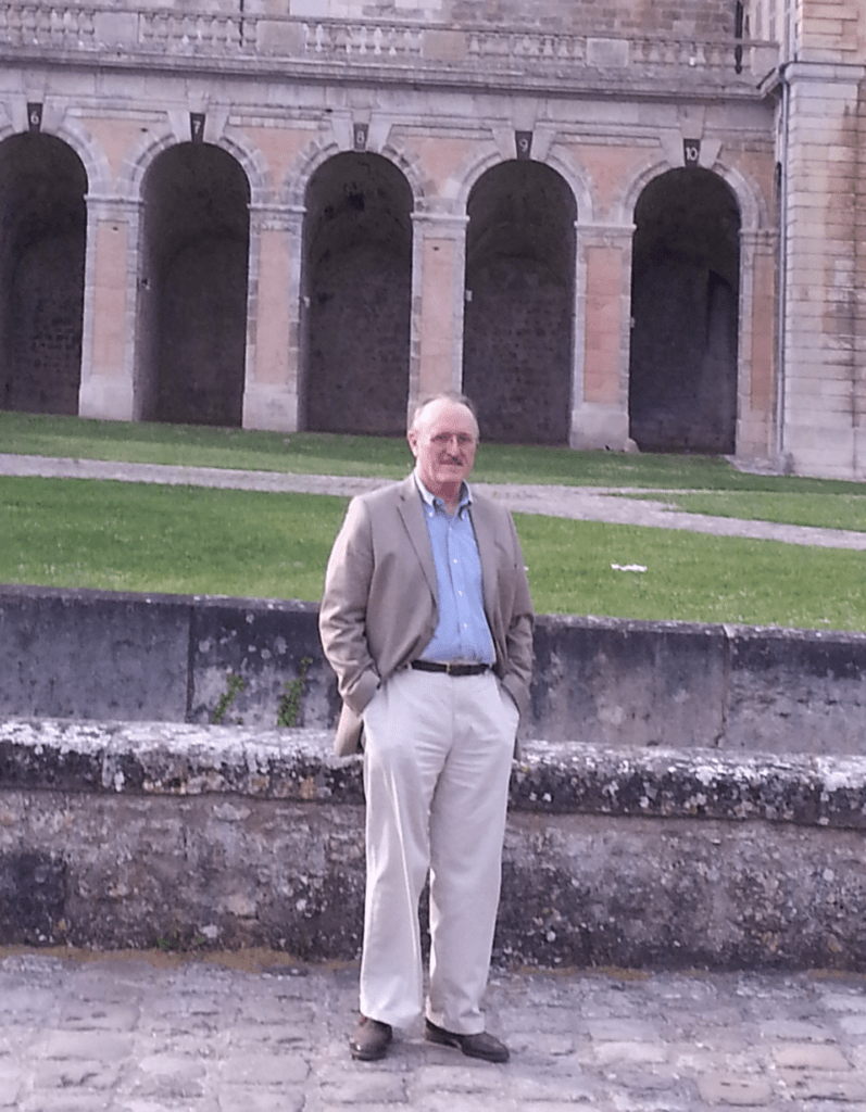 A man in a beige blazer and light blue shirt stands in front of an arched stone structure, surrounded by grass, suggesting a historical site.