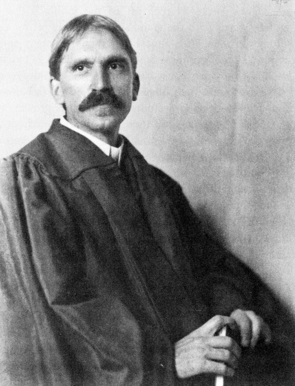 Portrait of a serious-looking man in a dark academic gown, seated, with a cane in hand, reflecting an intellectual demeanor.